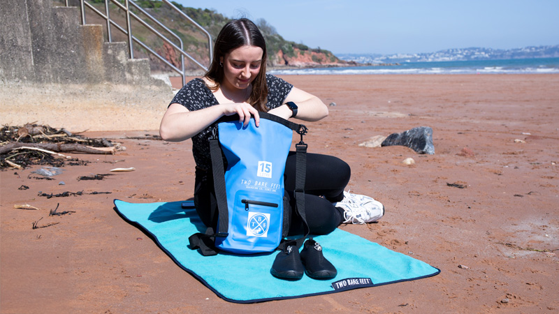 Woman sitting on the beach with a blue Two Bare Feet dry bag