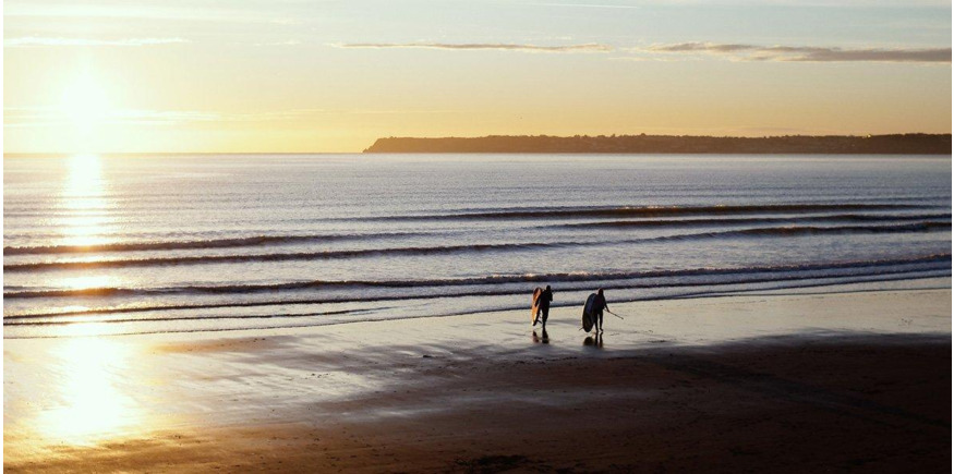 two paddleboarders walking down the beach towards the sea at sunrise 