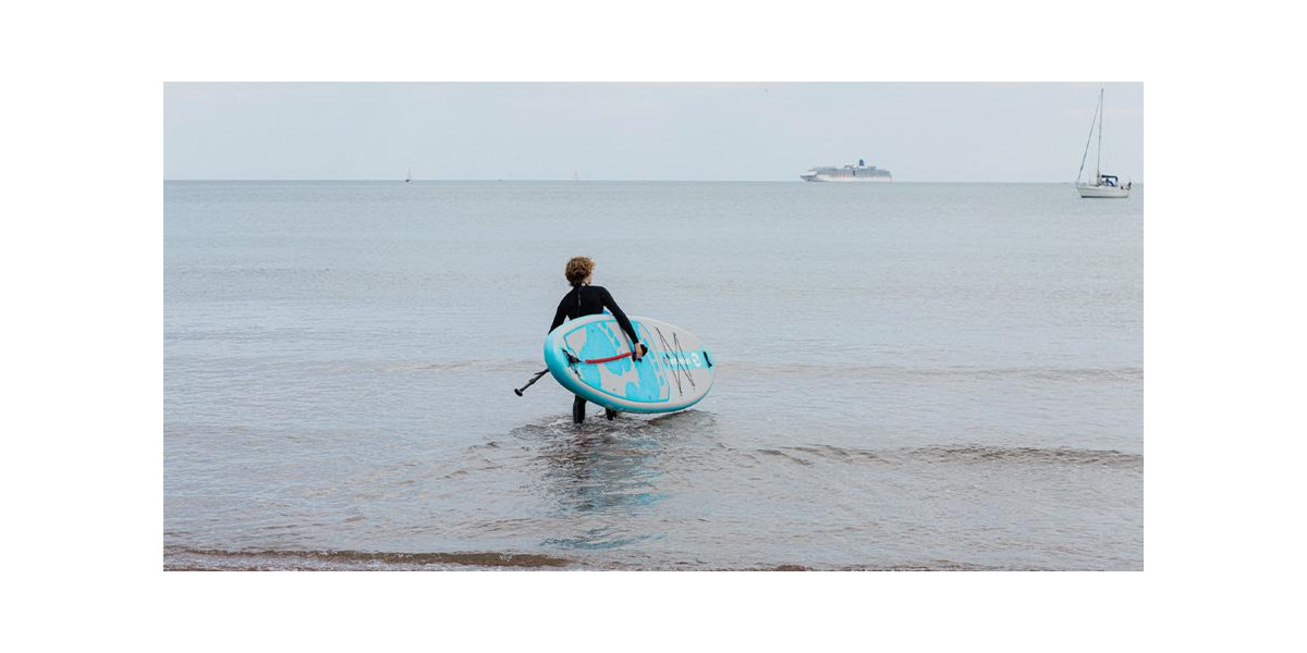 Paddleboarder in full body wetsuit carrying Two Bare Feet inflatable paddleboard into open water